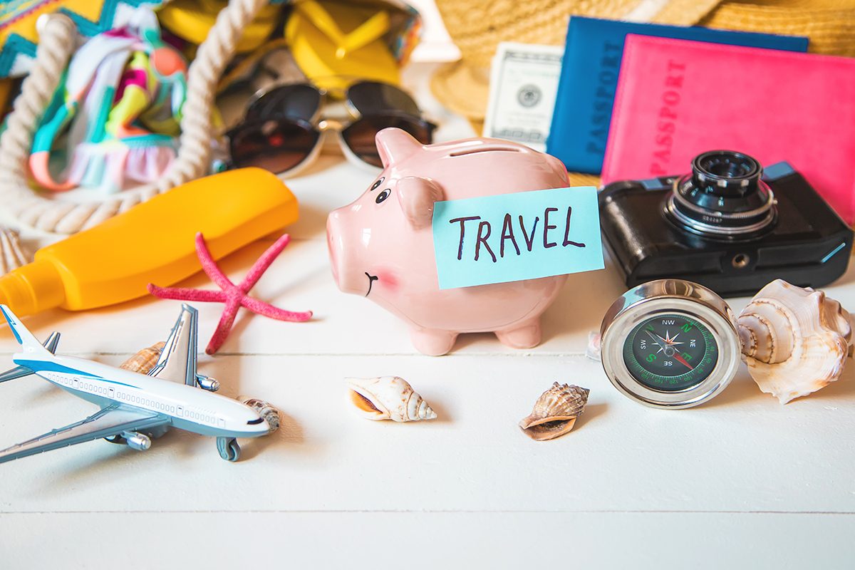 A collection of travel-related items, including a piggy bank with a sticky note that reads "TRAVEL" on it, a model plane, a camera, sun cream, passports, and shells, on a wooden surface