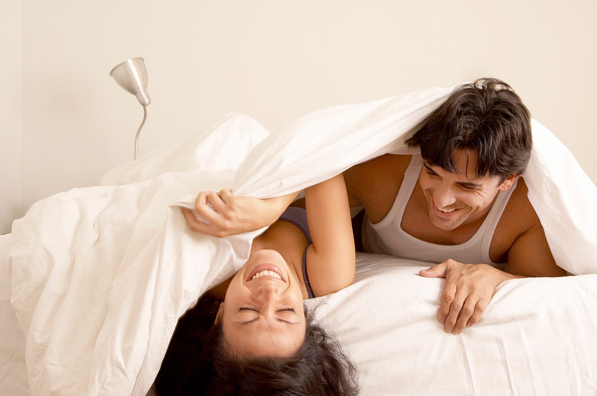 A couple lying on a bed under a white duvet, laughing