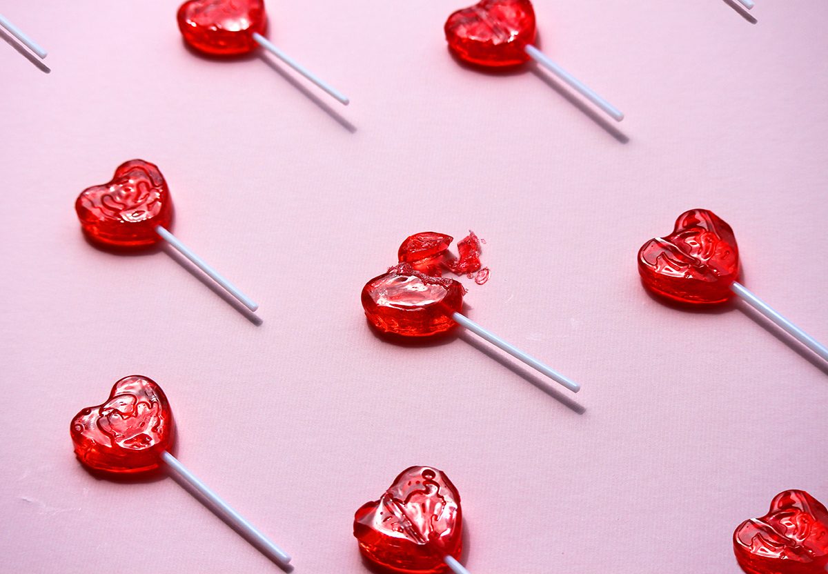 Red heart lollipops are arranged in diagonal lines on a pink background, with one broken
