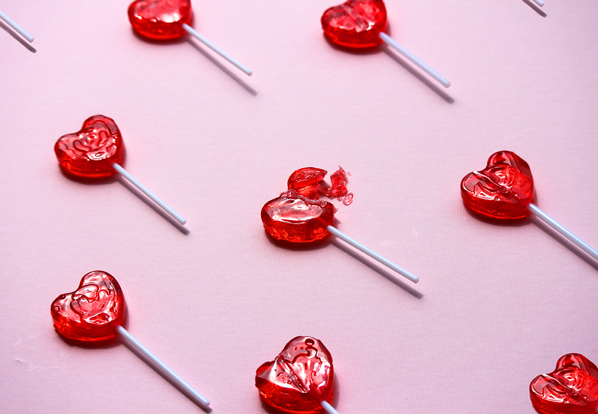 Red heart lollipops are arranged in diagonal lines on a pink background, with one broken