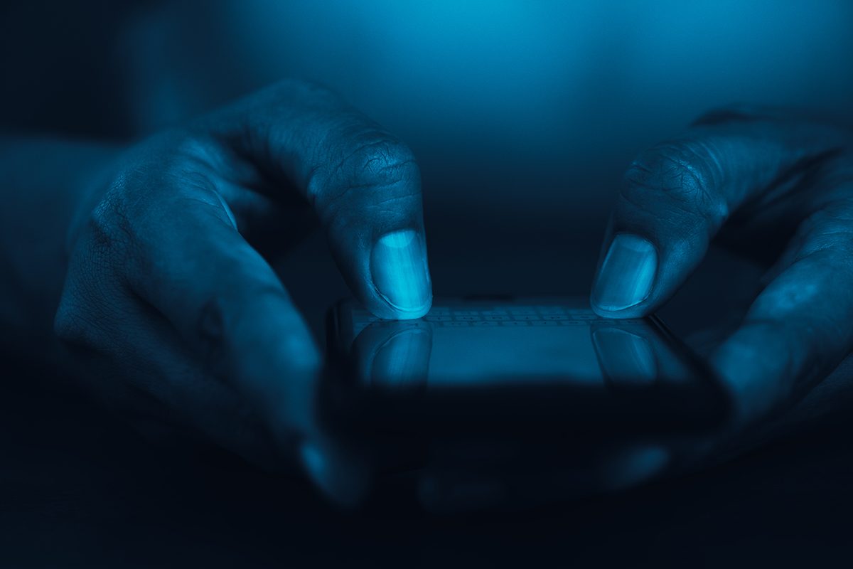 A close-up shot of hands typing on a smart phone. The photo has a blue tint. 