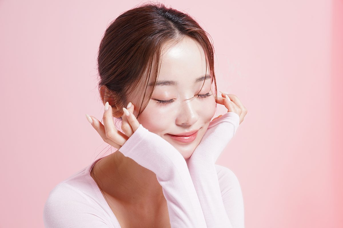 A woman has her eyes closed and is touching her face against a pink background