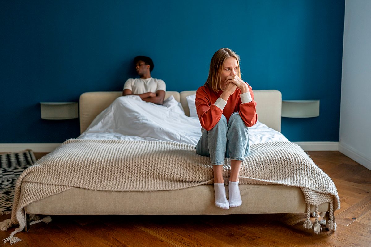 A sad woman sits on the edge of a bed while her unhappy partner looks away on the other side of the bed