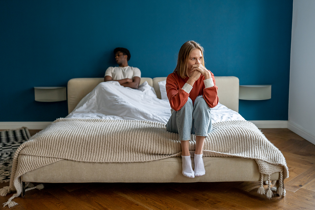 A sad woman sits on the edge of a bed while her unhappy partner looks away on the other side of the bed
