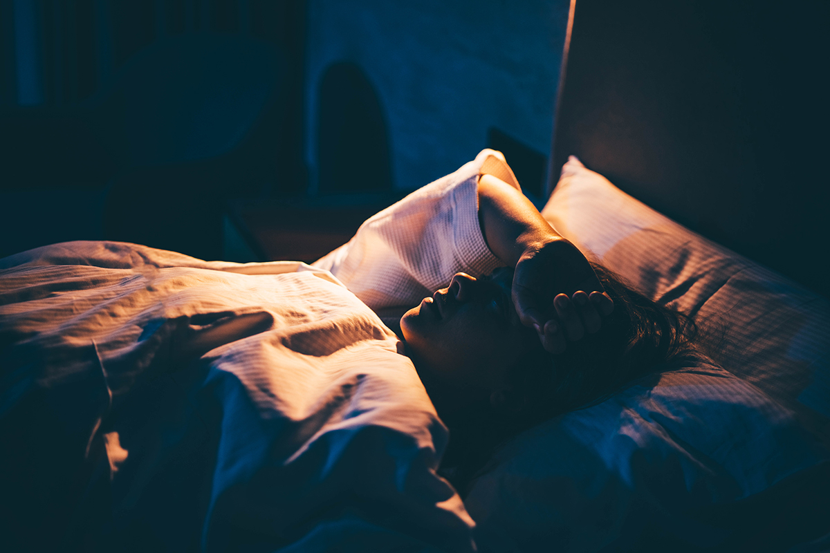 A woman lying on a bed in a dark room with her hand on her forehead