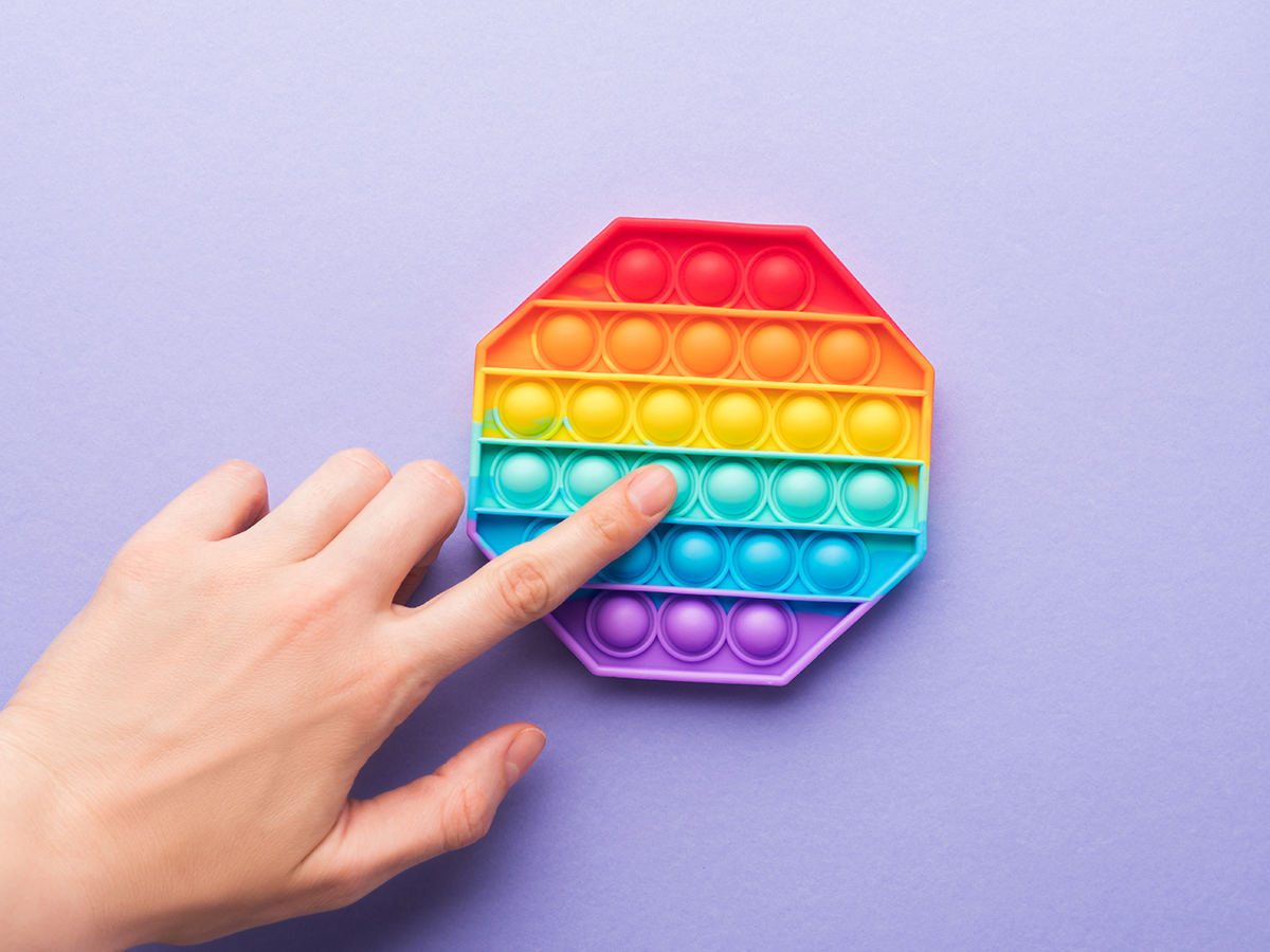 A hand playing with a rainbow-coloured Pop-It fidget toy against a purple background