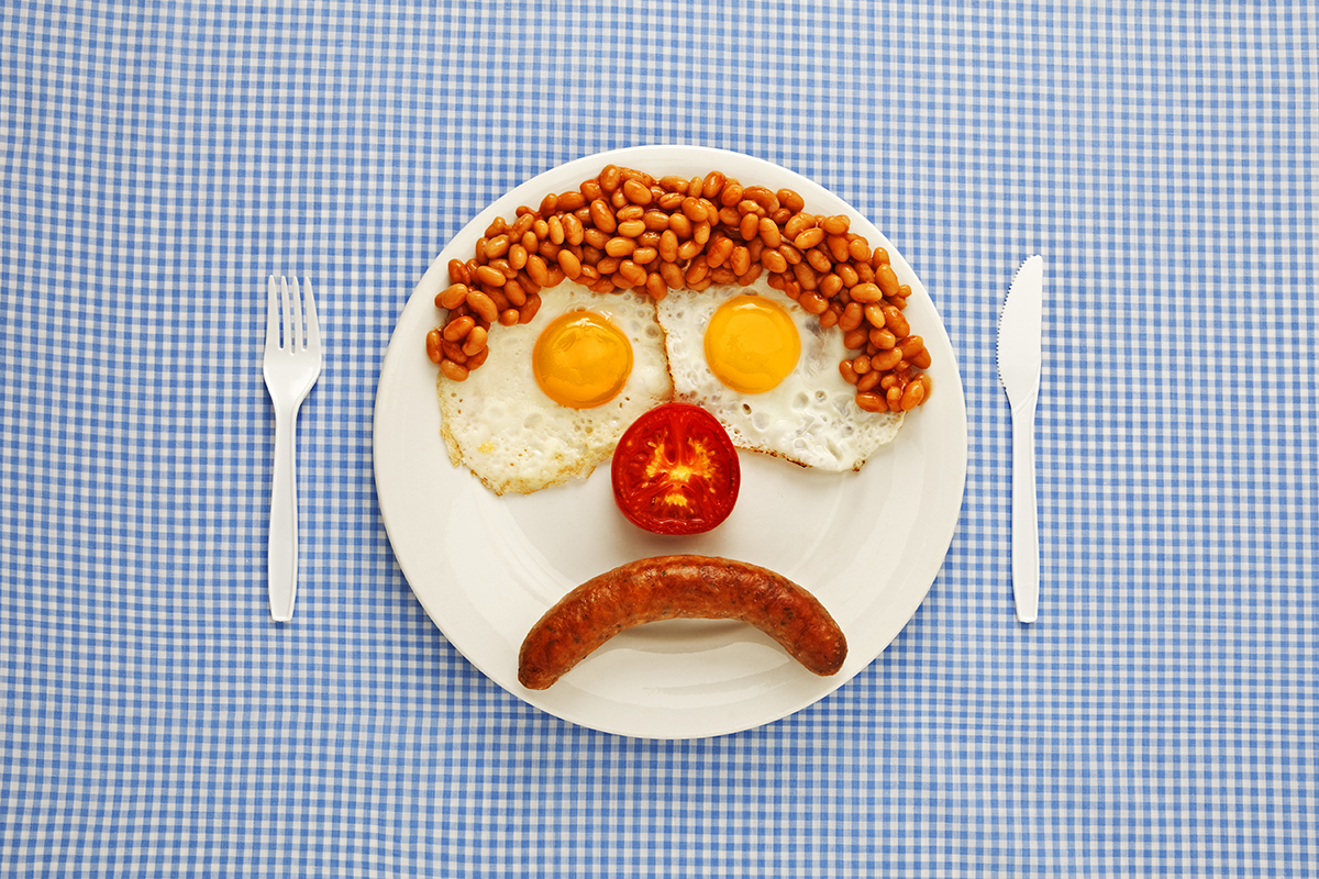 A fry up, arranged on the plate to resemble an unhappy face, sits on a blue gingham tablecloth.