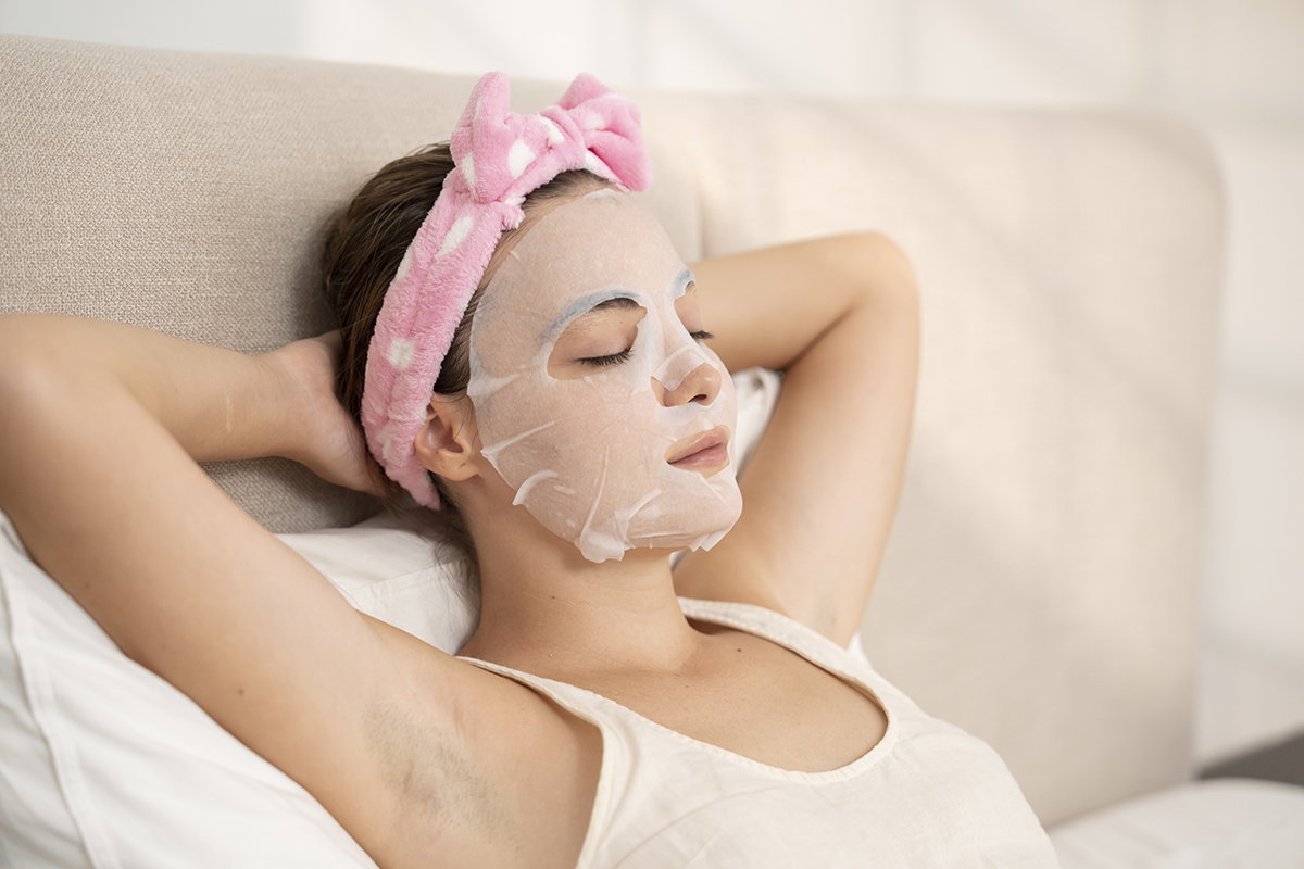 A woman wearing a sheet mask relaxes in bed with her eyes closed and hands behind her head.