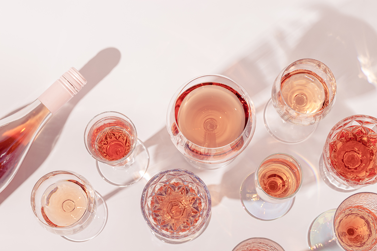 Different shaped wine glasses containing rose and a bottle of rose sit on a white background.