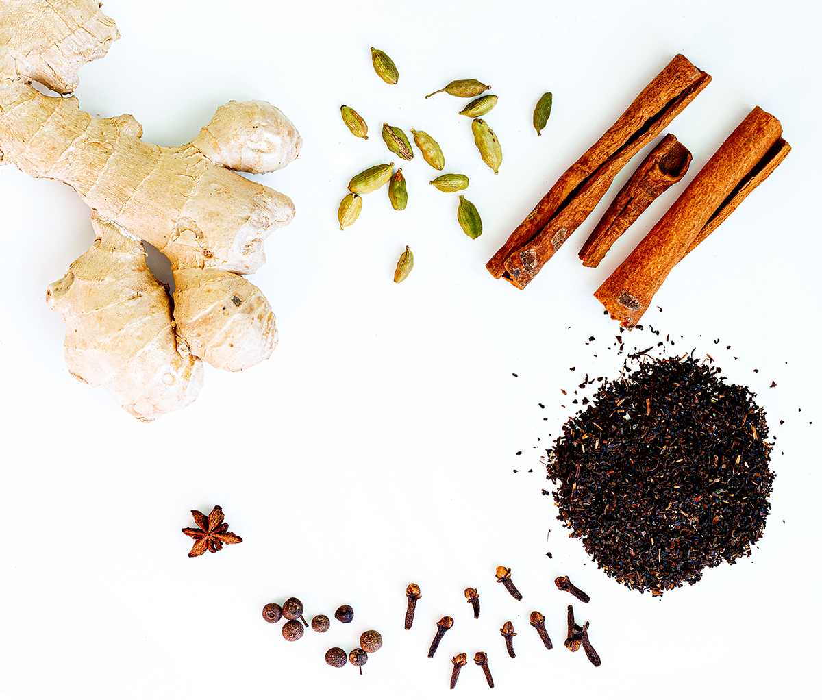 Ingredients for masala tea, including ginger, cinnamon sticks, cardamom, cloves and black pepper, are arranged on a white background.