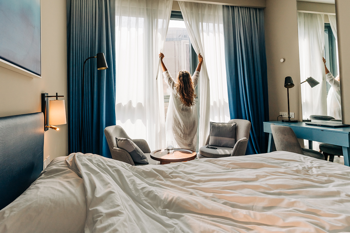 A woman in a robe stands at a window, pulling back white curtains to reveal daylight in a cosy hotel room. The room features a neatly made bed in the foreground.