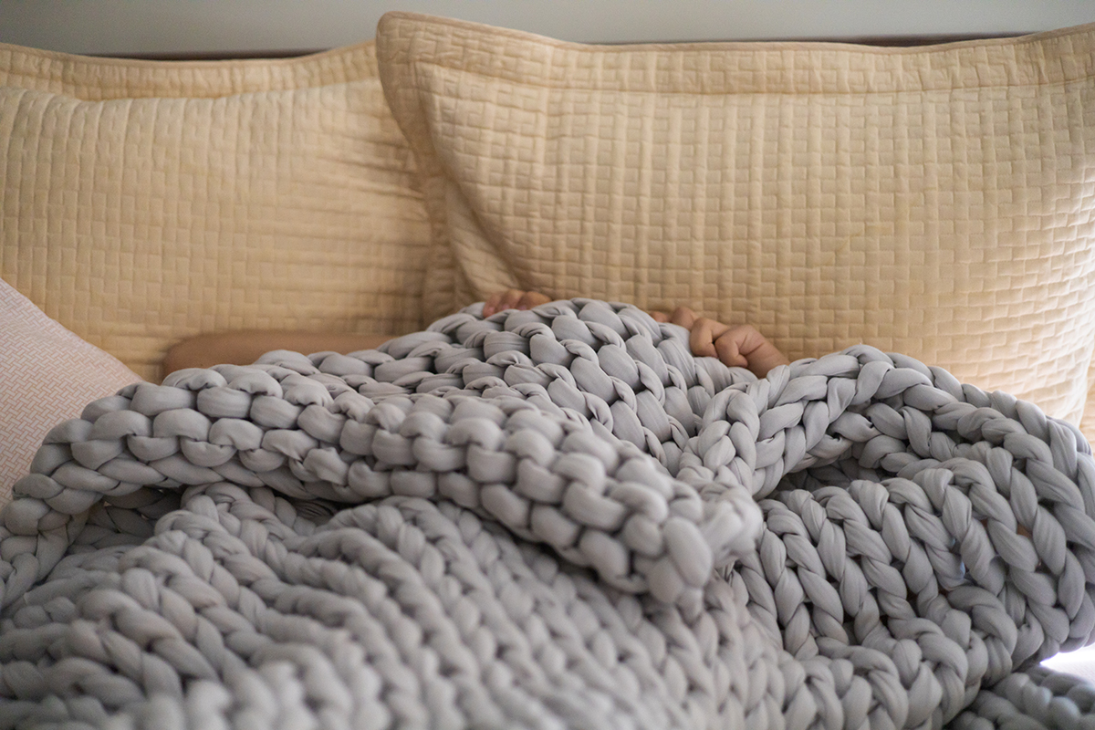 A person is tucked under a chunky grey knit blanket, with just a sliver of their face visible, resting against beige pillows.