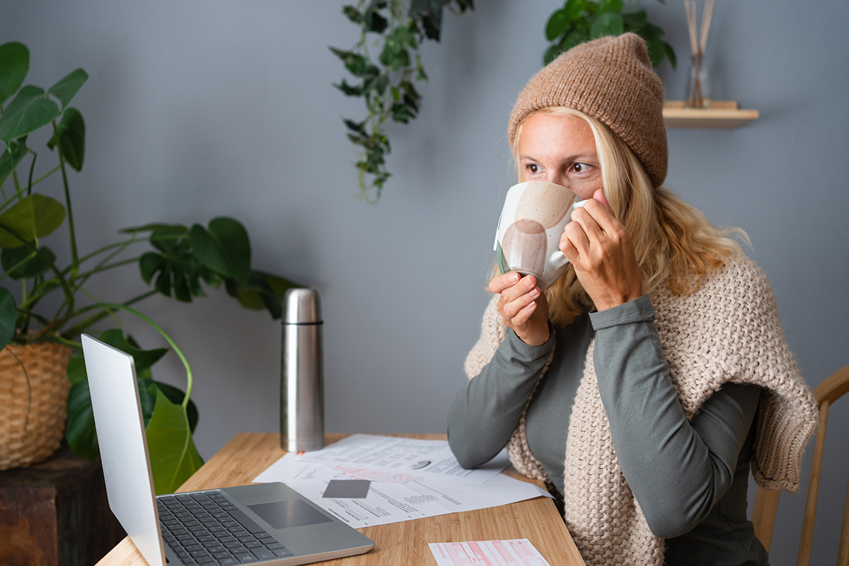 A woman wearing a knit hat and shawl sits at a wooden table, sipping from a mug while working on a laptop. 