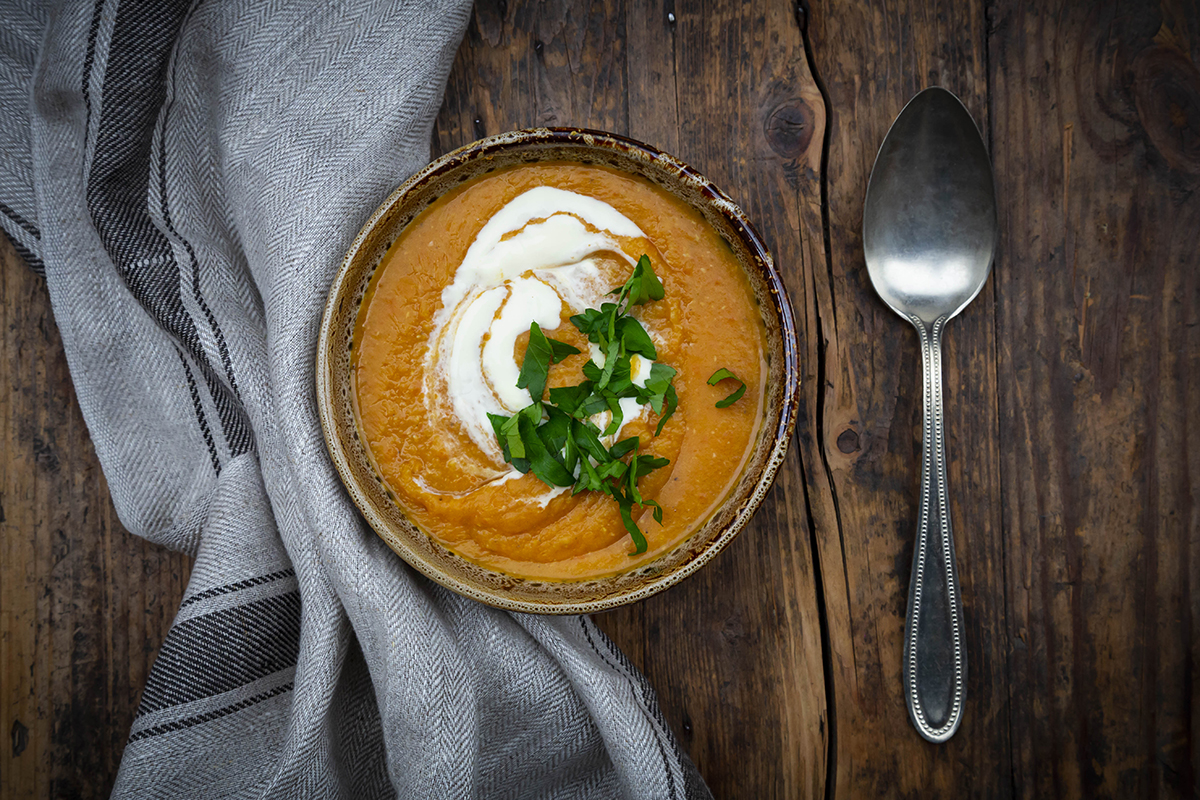 A bowl of red lentil soup garnished with a swirl of cream and fresh green herbs sits on a rustic wooden table, accompanied by a spoon and a folded gray cloth.