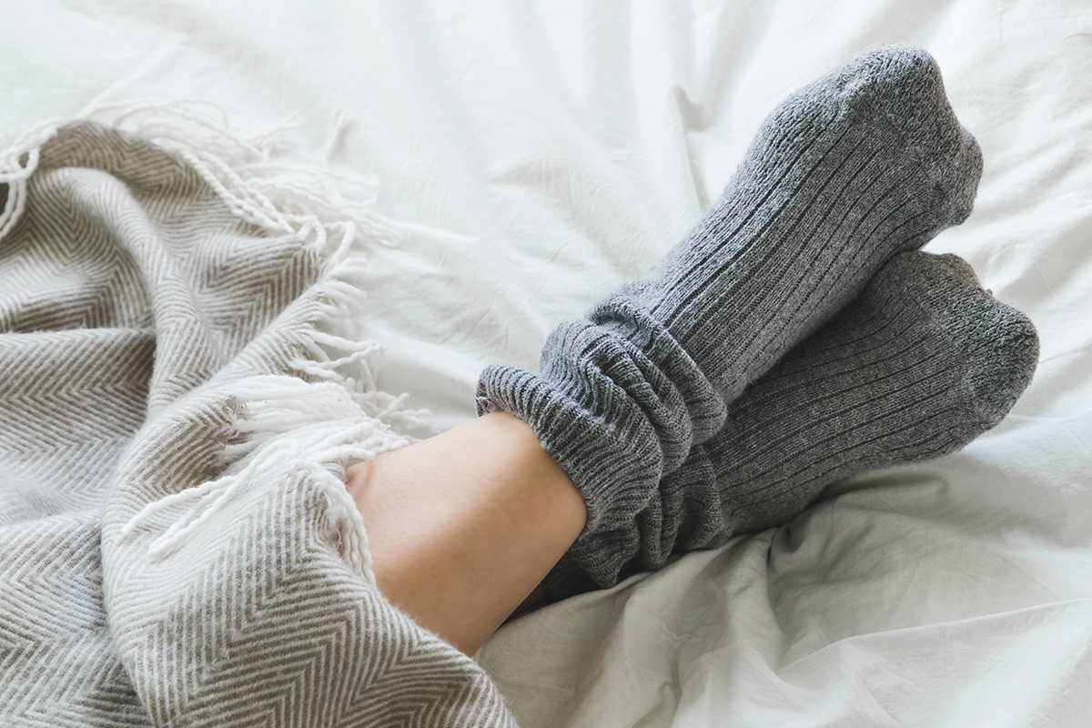 A pair of feet in grey socks are positioned on a bed under a cosy blanket.