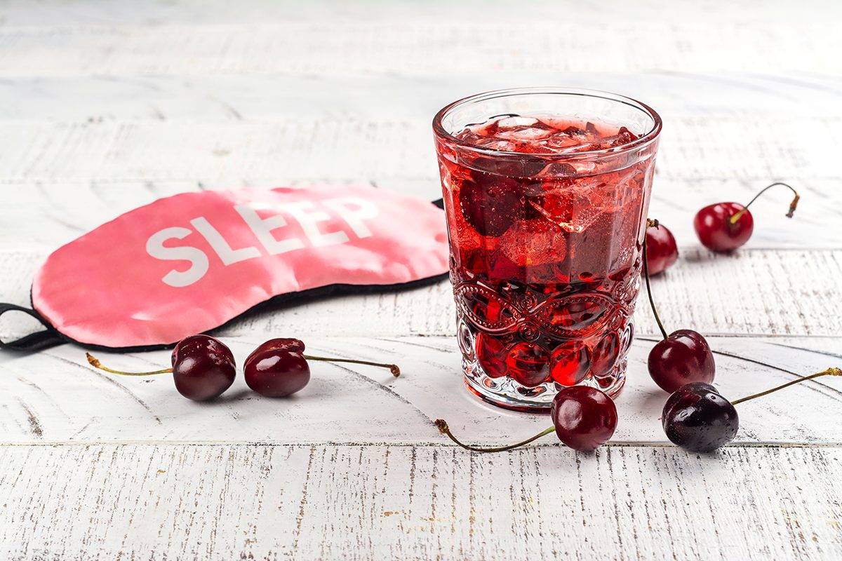 A glass of cherry juice, fresh cherries and a sleep mask with the word 'sleep' on it sit on a wooden surface