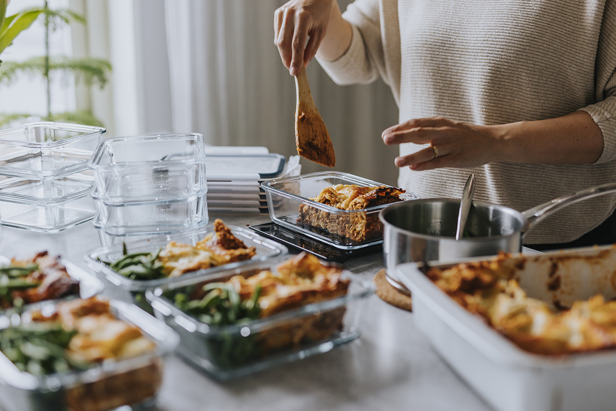 A person is preparing meals in a kitchen, using a wooden spoon to portion food into glass containers.