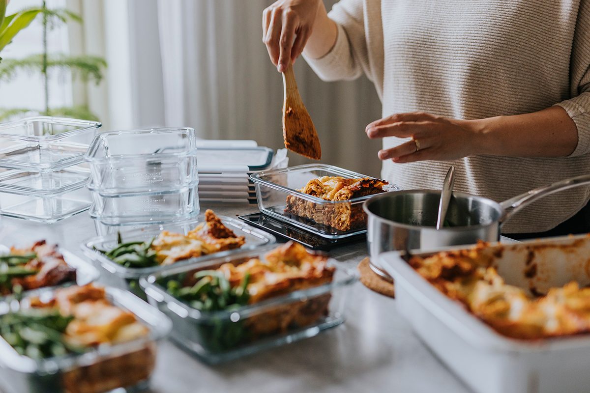 A person is preparing meals in a kitchen, using a wooden spoon to portion food into glass containers.