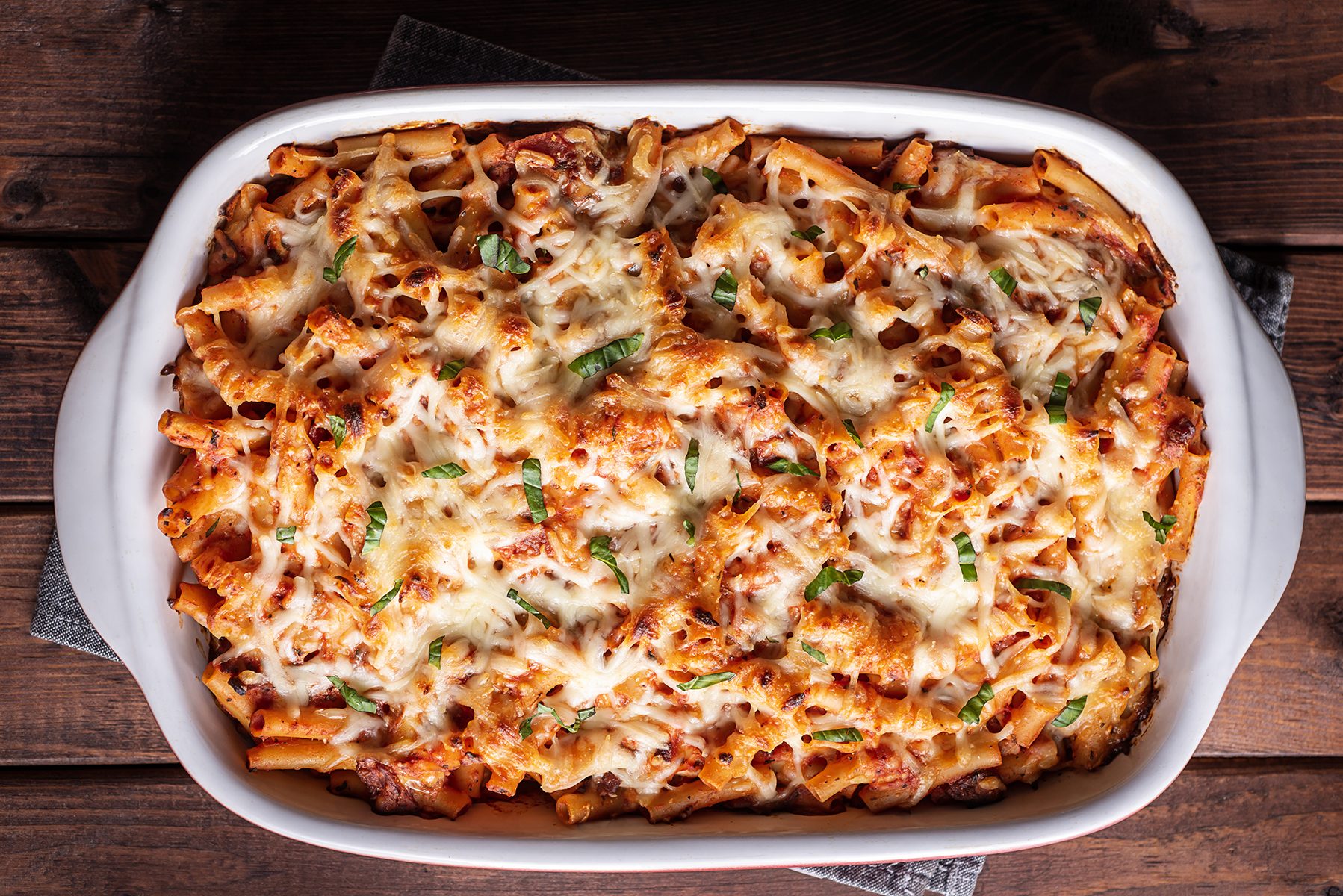 A baked pasta dish in a white casserole dish, topped with melted cheese and garnished with herbs, sits on a wooden table.