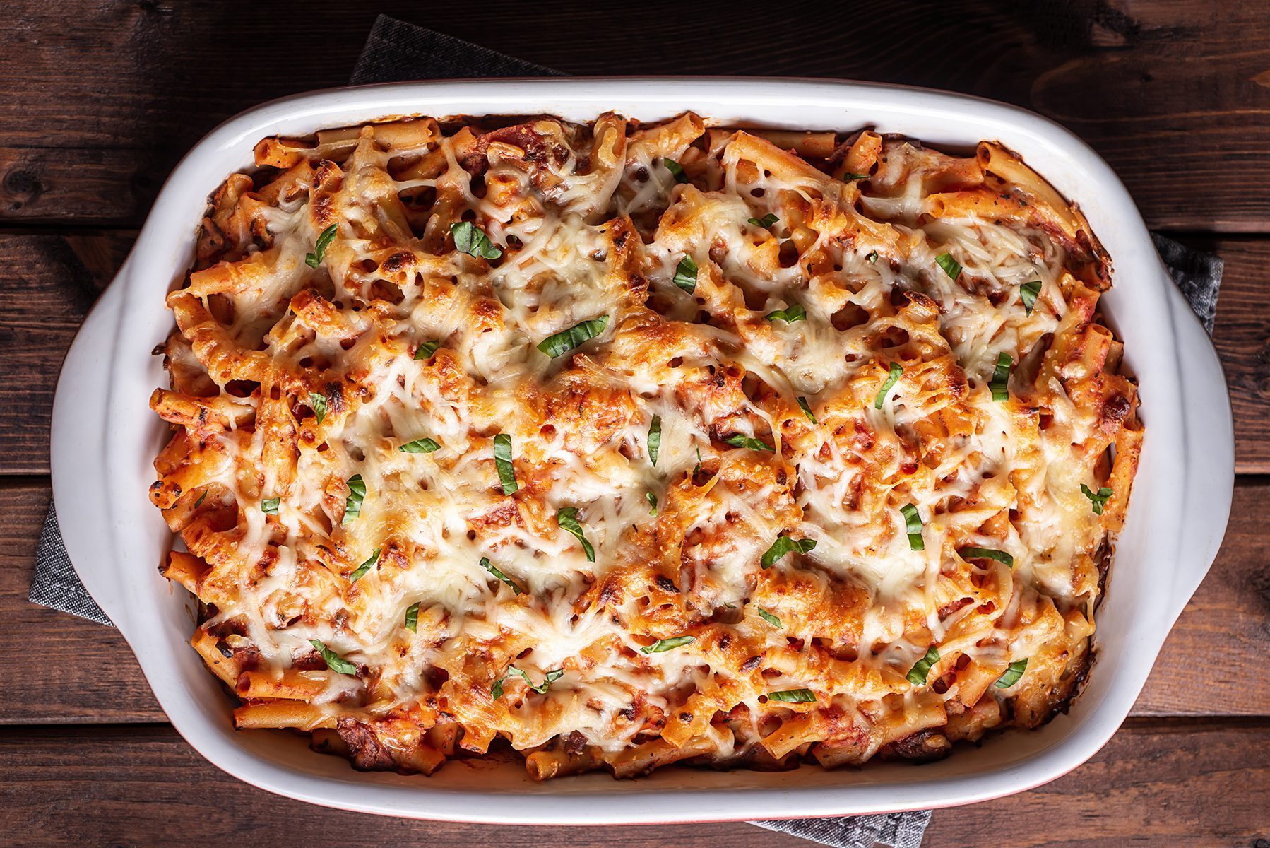 A baked pasta dish in a white casserole dish, topped with melted cheese and garnished with herbs, sits on a wooden table.