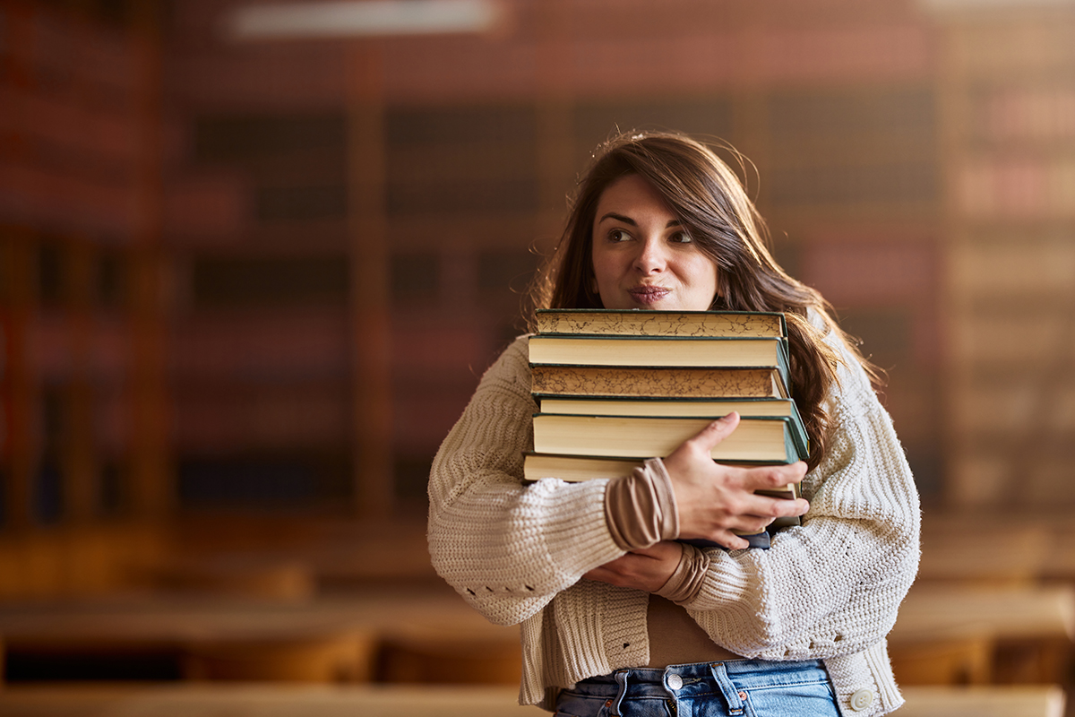 A student carries a pile of books in front of a blurred background of a library.