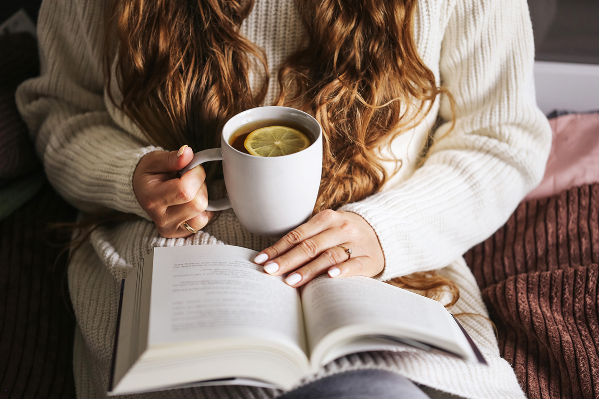 A cropped shot of a woman holding a mug of tea with a book in her lap.