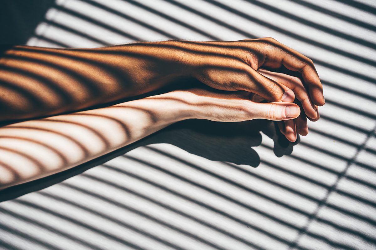 Two arms are stretched out across a bed, their hands intertwined, with the shadows of blinds falling over them.