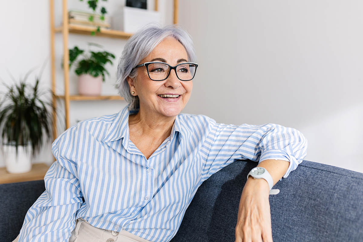 A mature woman wearing a pinstriped blue and white shirt relaxes on a sofa, smiling.