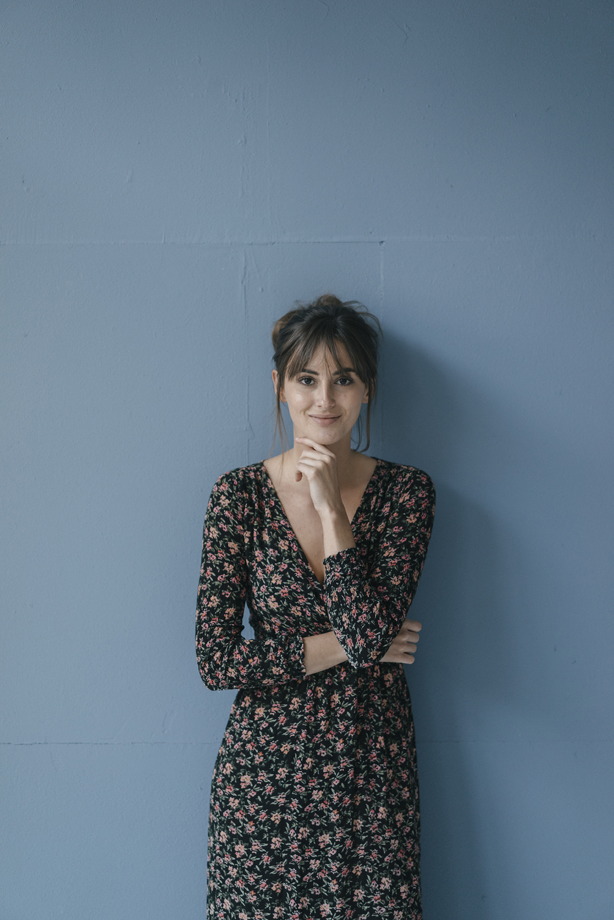 A woman wearing a long floral dress leans against a blue wall.