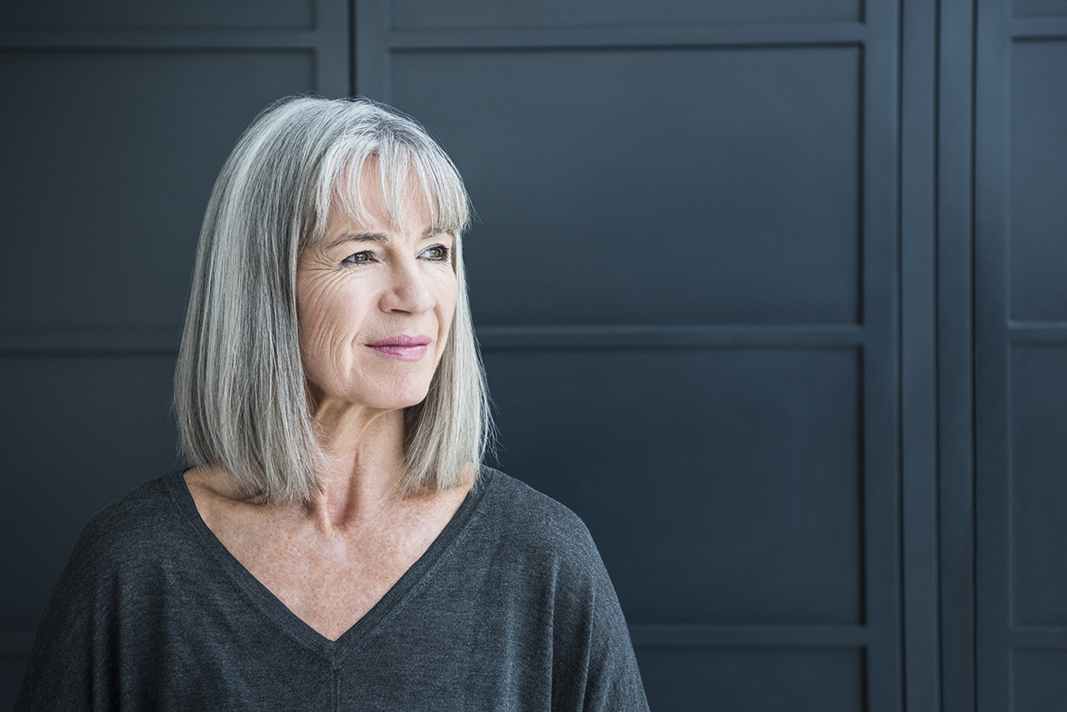 A mature woman with a grey bob and fringe stands in front of a dark grey background.