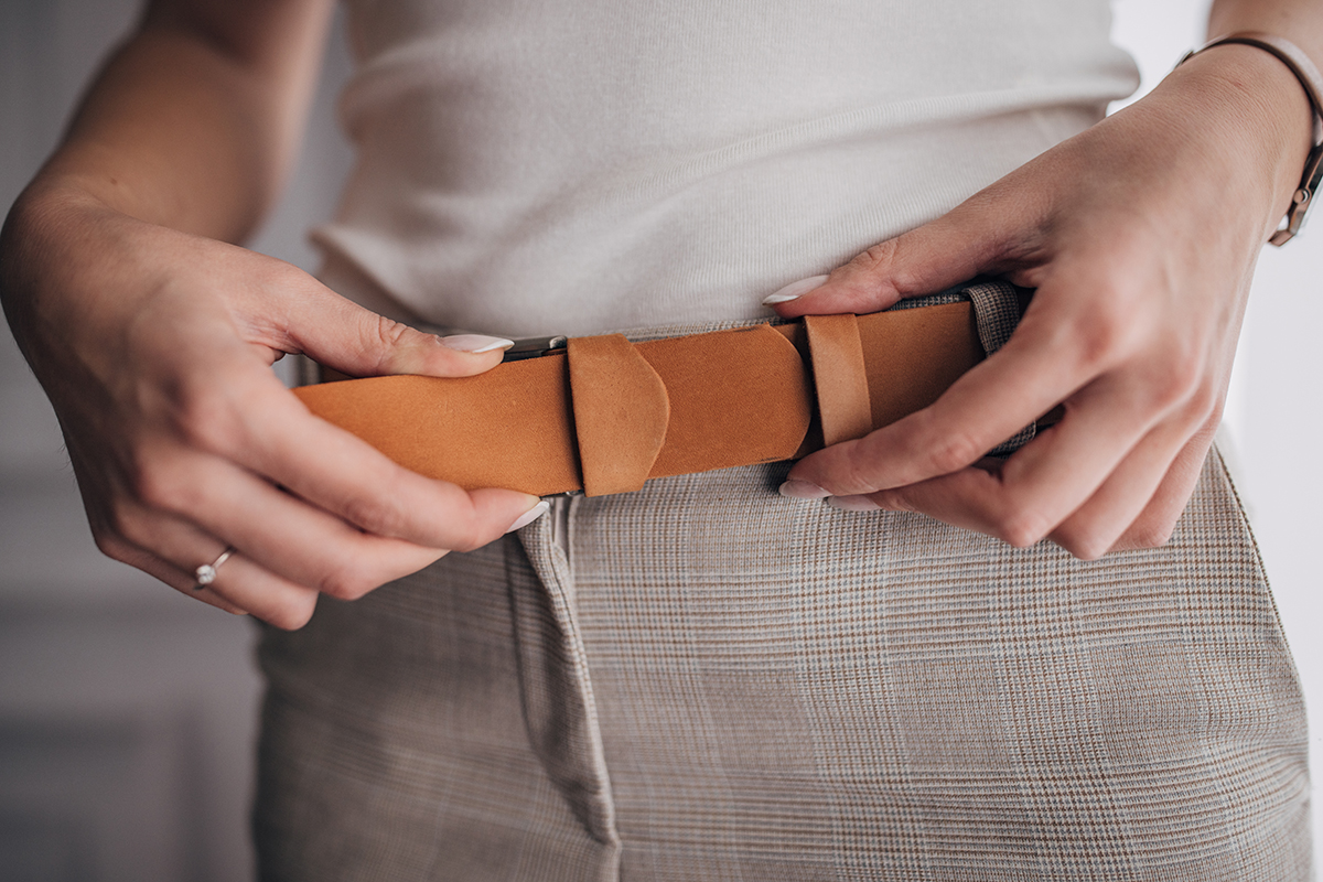 A close up shot of hands fastening a light brown leather belt.