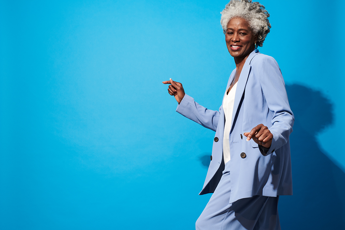 A middle-aged woman wearing a light blue suit is dancing in front of a blue background.