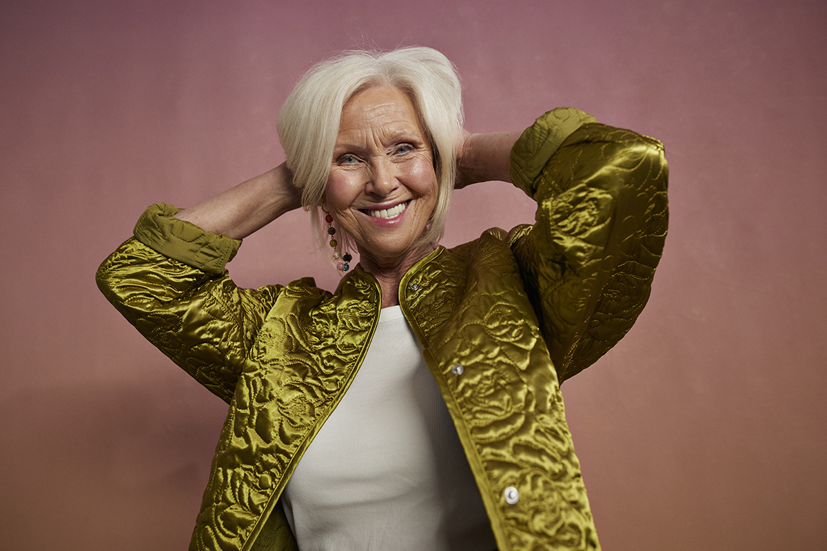 A mature woman wearing a shiny green jacket poses with her hands behind her head in front of a pink background.