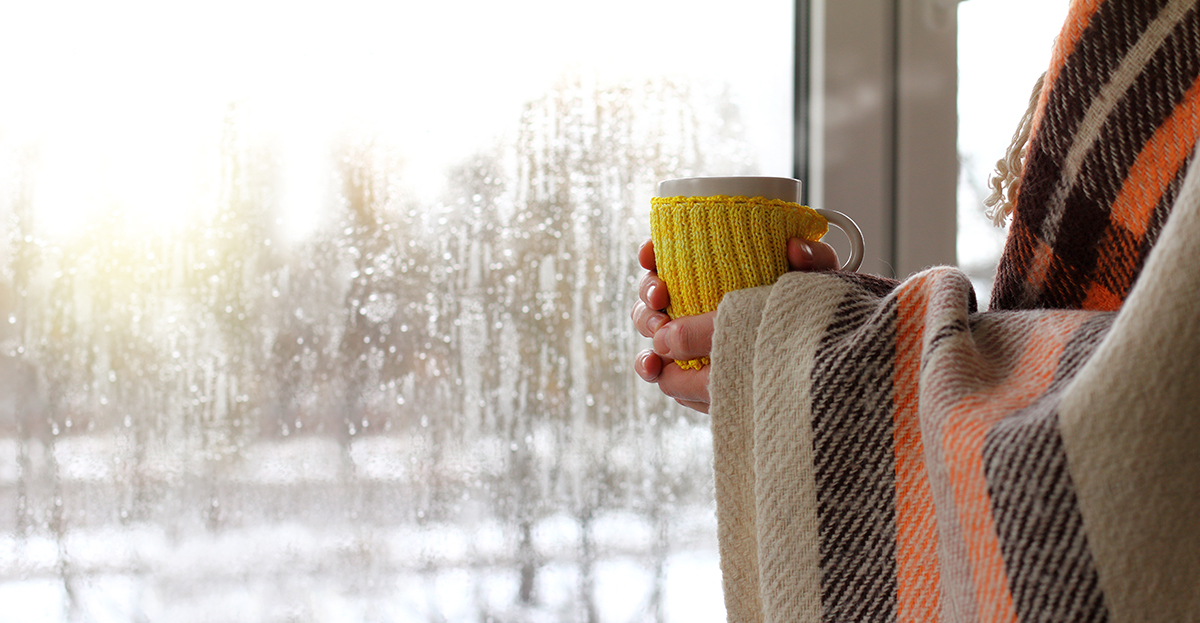 A person wrapped in a blanket, holding a mug stands next to a window.