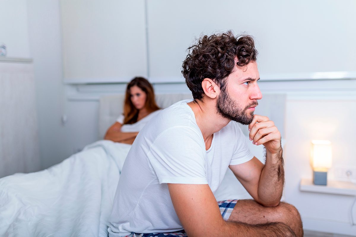 A man sits on a bed thinking, with his girlfriend, who has her arms crossed, in the background
