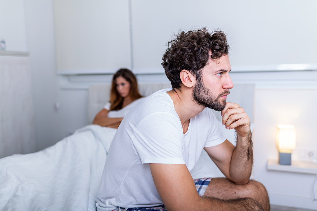 A man sits on a bed thinking, with his girlfriend, who has her arms crossed, in the background