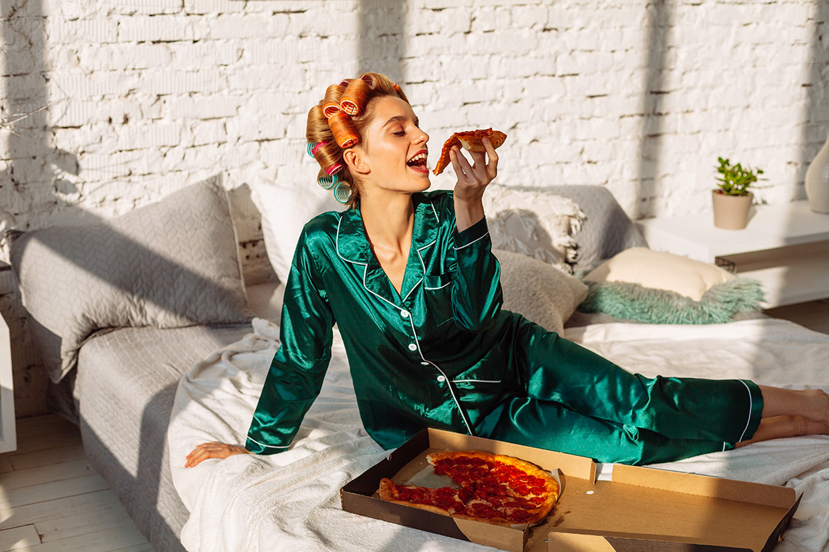 A woman wearing silk pyjamas eating pizza in bed