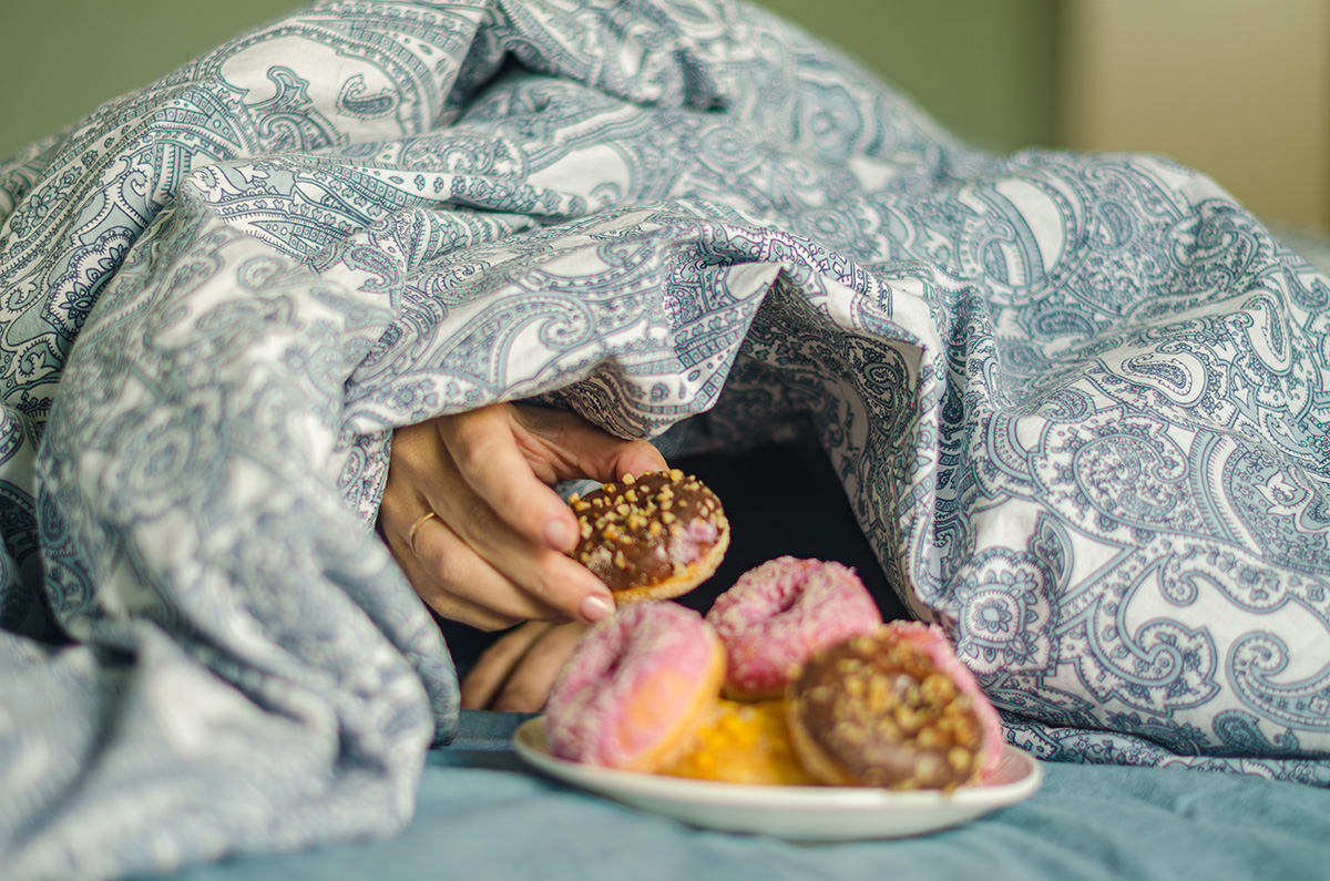 A hand reaches out from under a duvet, picking a doughnut of a plate