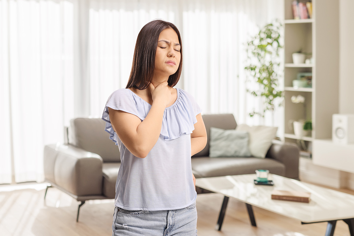 A woman stands in a living room, holding her throat in pain