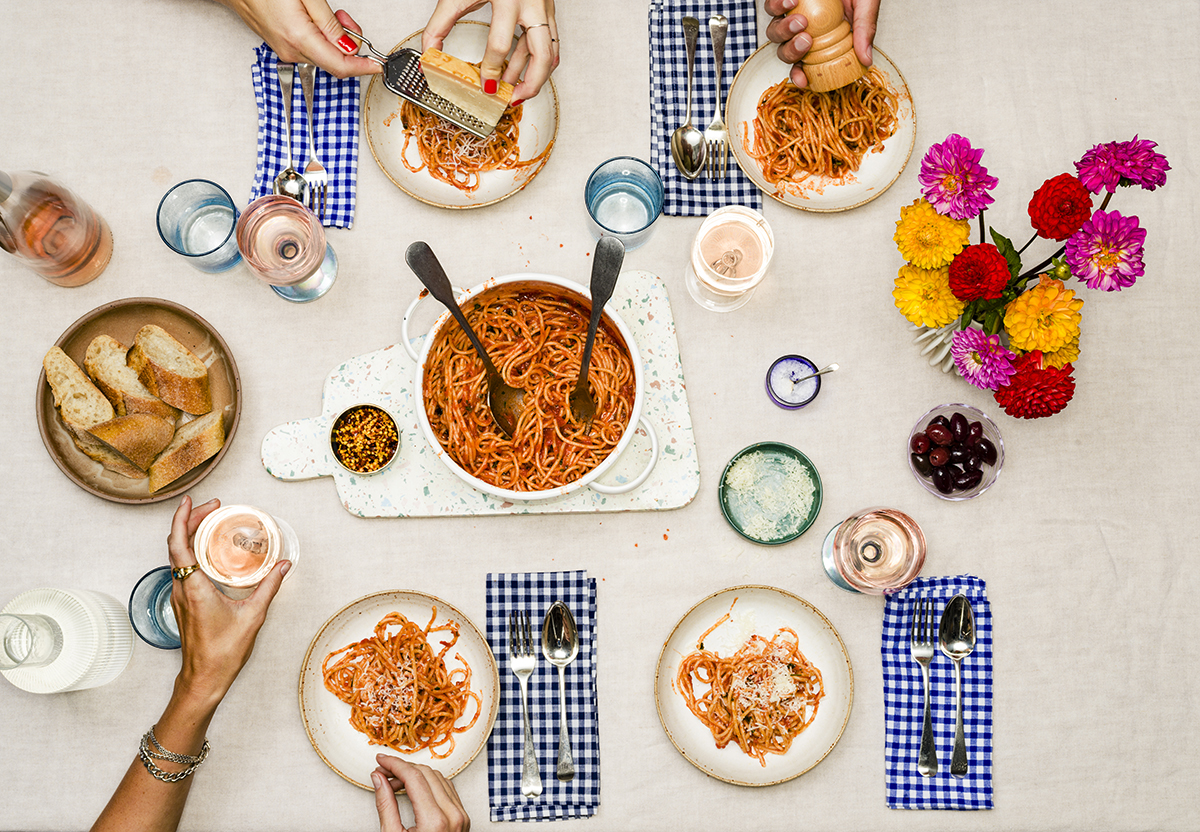 An overhead shot of friends eating together at the dinner table. The table features a bowl of spaghetti, bread, glasses of wine and a vase of fresh flowers