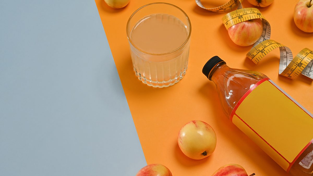 A glass and bottle containing apple cider vinegar sit on a grey and orange surface, surrounded by apples and a measuring tape
