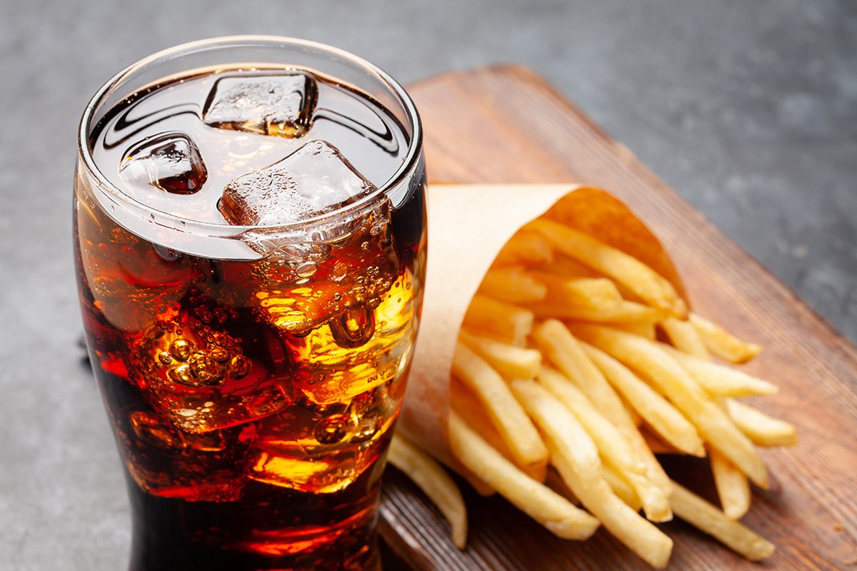 A glass filled with cola and ice and a portion of french fries sit on a wooden board