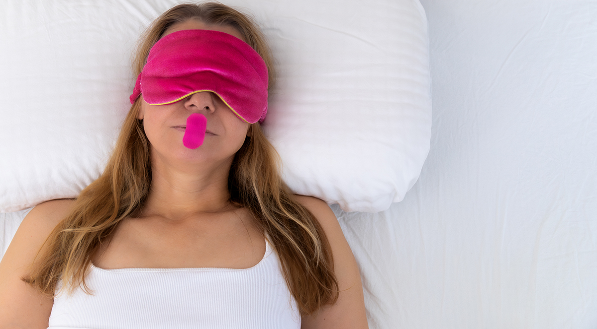 A woman wearing a pink eye mask and mouth tape sleeps in bed
