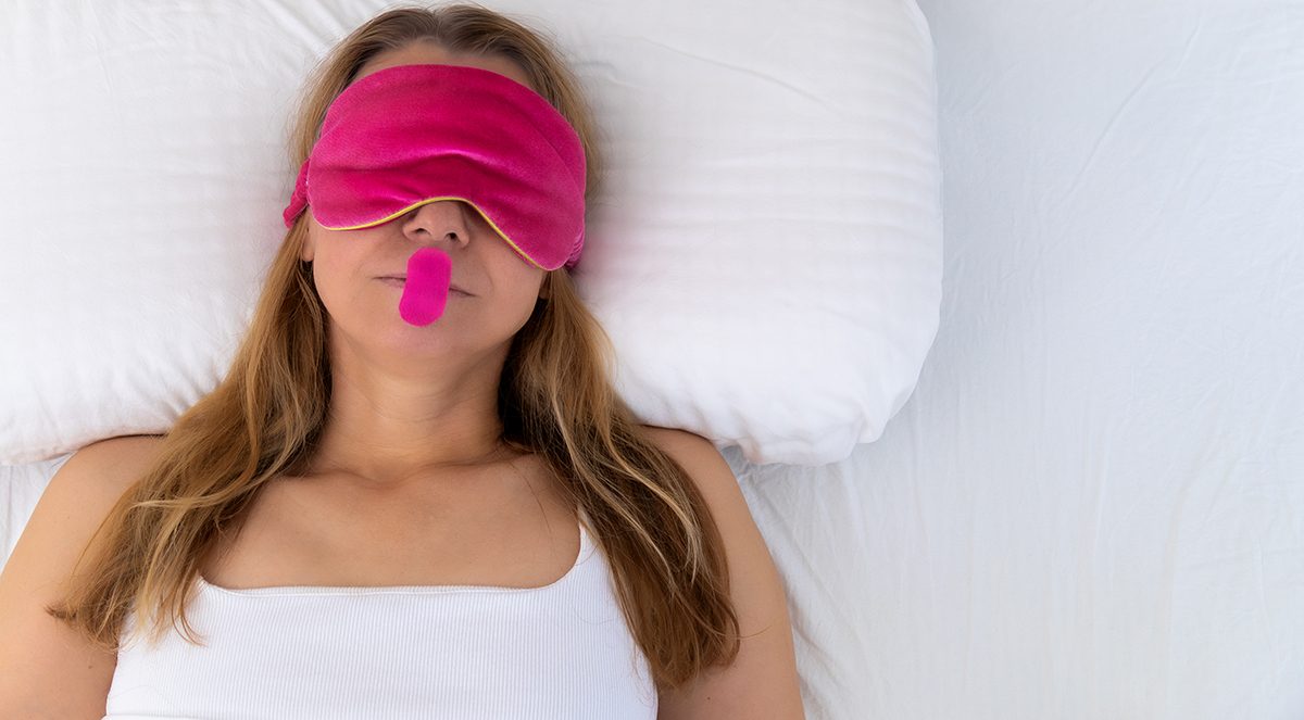 A woman wearing a pink eye mask and mouth tape sleeps in bed