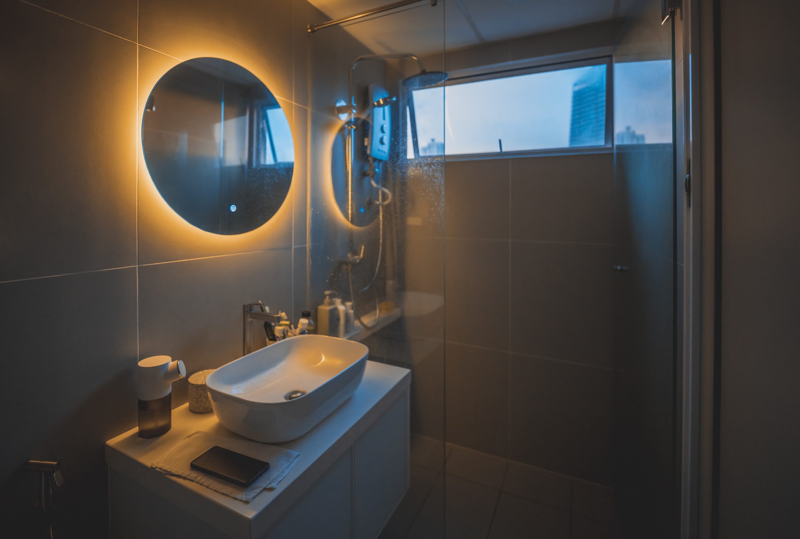 A modern bathroom features a glowing round mirror above a sleek white basin on a countertop.