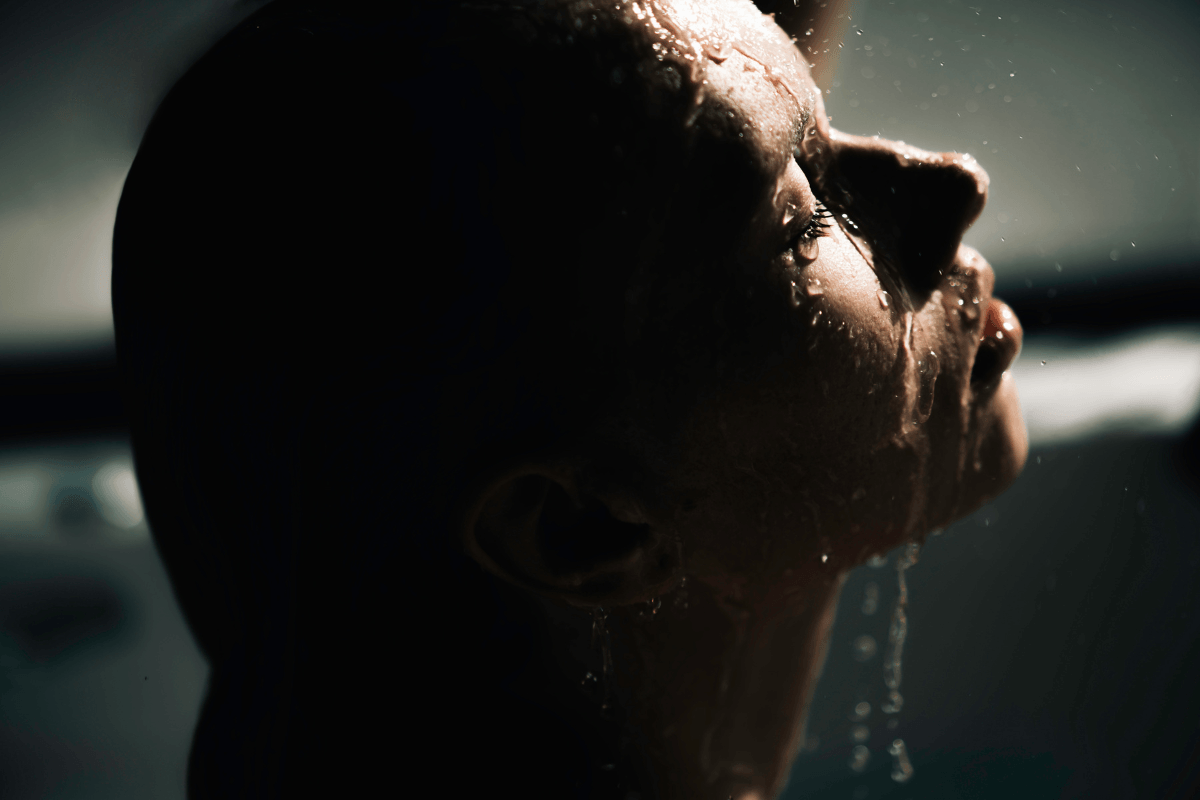A woman is standing under running water in a dimly lit setting. Droplets are cascading down her face.
