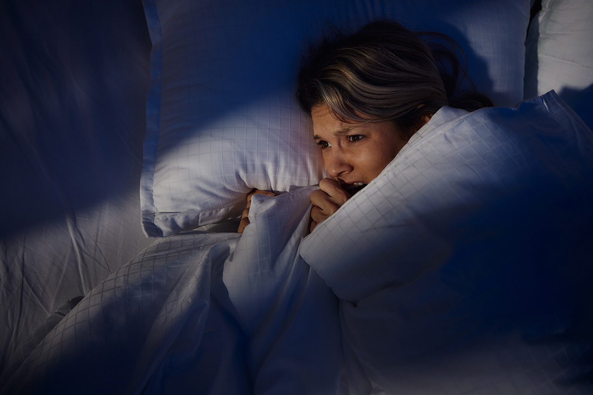 A scared-looking woman is lying in a bed, with the duvet pulled up around her, at night
