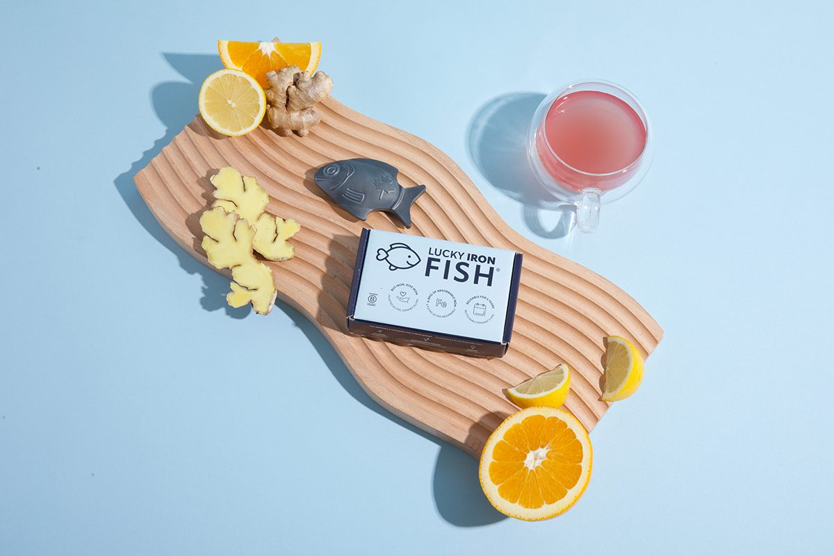 The Lucky Iron Fish cooking tool on a wooden board next to slices of citrus fruits, ginger, and a pink drink, set against a blue background