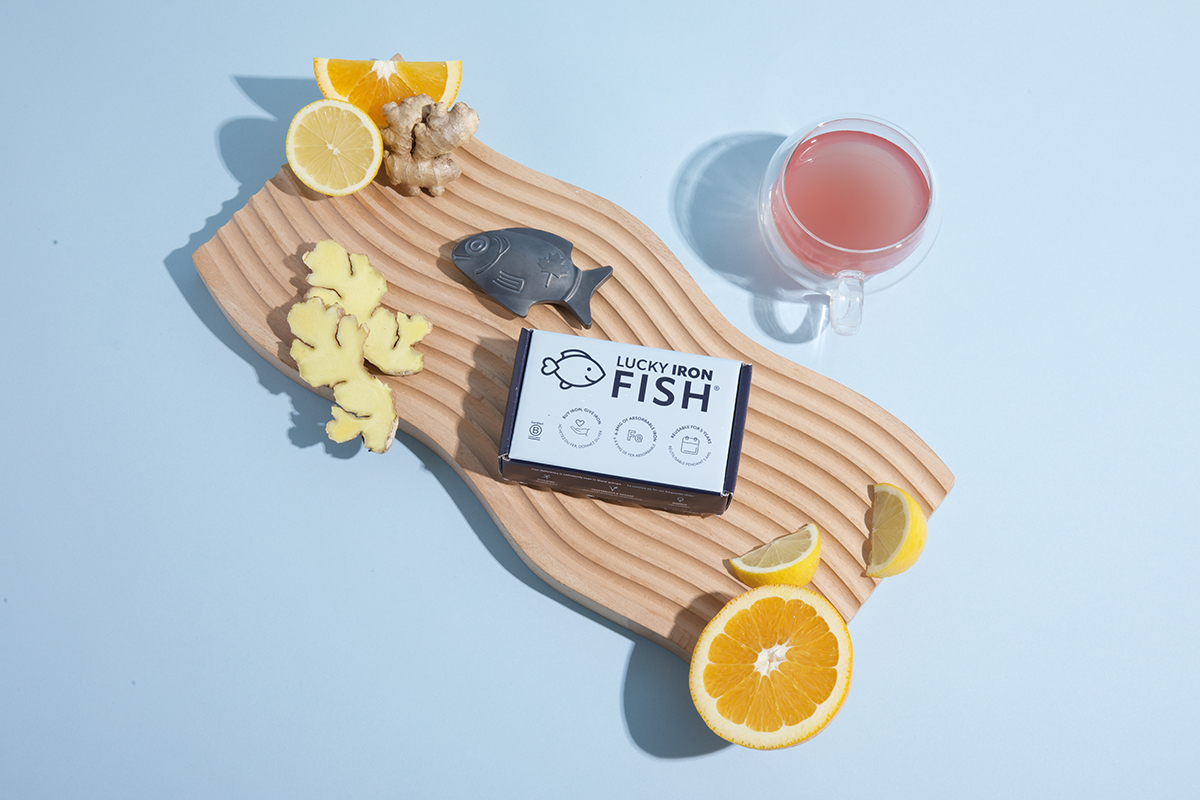 The Lucky Iron Fish cooking tool on a wooden board next to slices of citrus fruits, ginger, and a pink drink, set against a blue background
