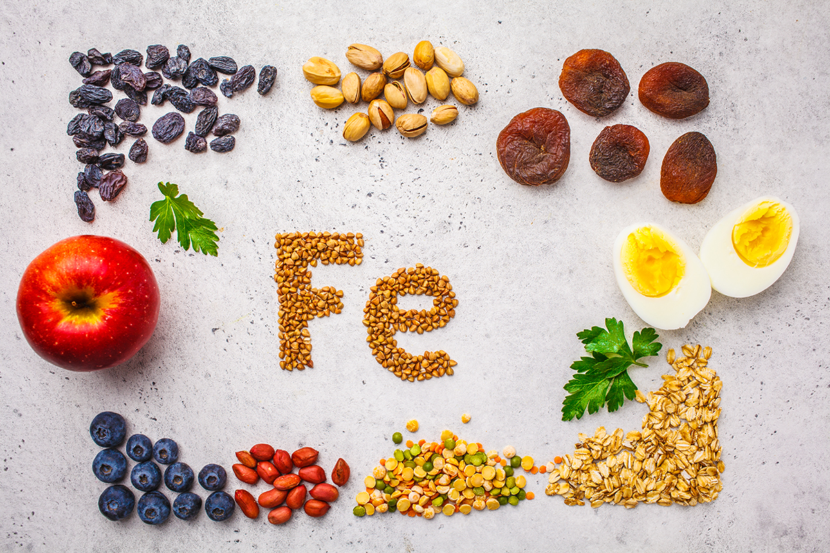 An assortment of iron-rich foods, including fruits, nuts, lentils and eggs, are displayed on a surface with buckwheat, that has been arranged to read "Fe", in the centre